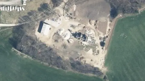 An aerial image showing what looks like a farm. There are various buildings on the land which is surrounded by greener ground. The trees left around the outskirts cast shadows on the floor.