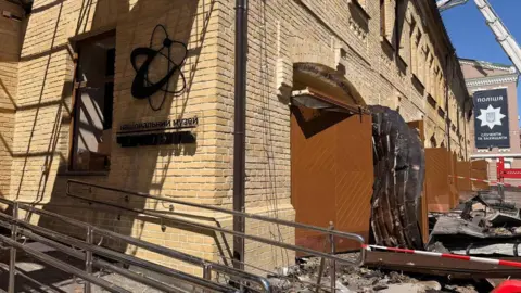 Ministry of Internal Affairs of Ukraine Brick museum exterior damaged, debris outside, emergency equipment and police sign visible.