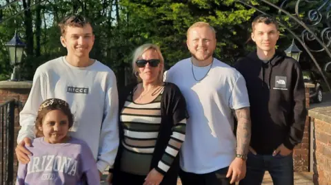 Brain Tumour Research A family - three young men and a young girl, are standing around Stephanie, smiling at the camera. The su is shining and Stephanie is wearing sunglasses.