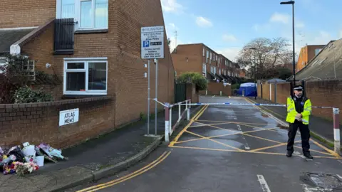 A crime scene, police tape and a police officer wearing high viz clothing.