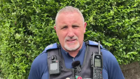 A PCSO with grey hair and a grey beard. He is wearing a police uniform with a radio on the shoulder of a stab vest.