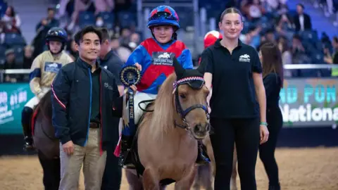 Hong Kong International Horse Show Harriet in blue and red riding costume sitting on her light brown Shetland pony, standing next to race officials holding her winners ribbon
