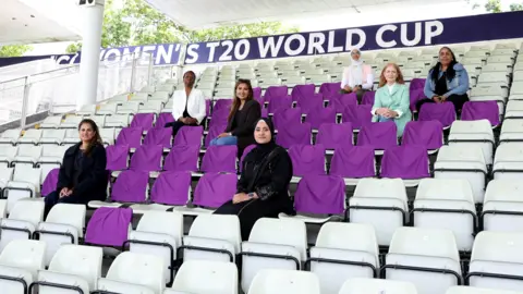 ECB Seven individuals are seated across several rows in a covered cricket stadium stand, spaced apart among pale-green plastic seats, many of which are draped with purple covers. In the foreground, one person dressed in black clothing and a headscarf sits centered, facing the camera. Other individuals are positioned higher up across the rows, wearing a mix of outfits including a white blazer, a dark jacket, a denim jacket, and a light green coat. A large banner behind them reads “Women’s T20 World Cup,” and a section marker “26” is visible on a pillar to the left. The stand is open at the sides, with greenery visible outside.
