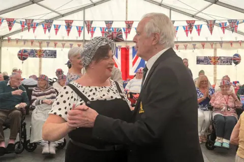A man with grey hair and a woman with dungarees and a headscarf dance together