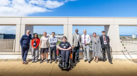 Creo Comms A review panel created from the city's community organisations has been shortlisting the best name suggestions for the new footbridge. The group is standing on part of the new bridge, with a woman in a wheelchair out in front.