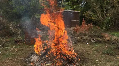 CDDFRS A bonfire is alight in a wooded area with a hut a few metres behind it. Next to the fire is a metal brazier 