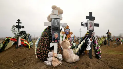 Goktay Koraltan/BBC A number of wooden crosses within a cemetery. One on the right is the grave of Tetiana Tarasevych and displays her photos. The cross on the left is the grave of Sophiia Buhayova and her son Adam. Both graves are covered in artificial flowers and teddy bears. 
