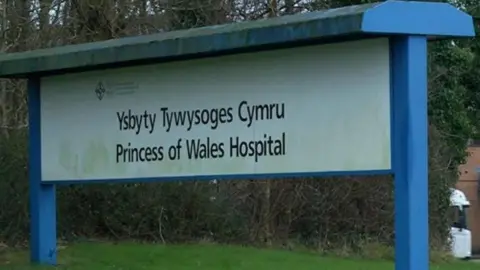 the sign in front of a forested area on the Princess of Wales hospital grounds. the hospital names is written in welsh and english on a white background framed by  blue sign posts 