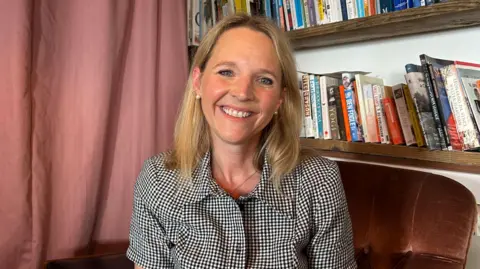 Kate has blonde hair and is wearing a black and white checked shirt. She is sat in front of a bookcase