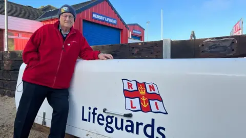 Lyndon Bevan, chairman of Visit Great Yarmouth, standing near the RNLI Lifeguards equipment store, which is white and bears the RNLI flag logo. Mr Bevan is wearing black trousers, a red jacket and a Save Hemsby Coastline beanie hat. He is standing outside the Hemsby Lifeboat Station.