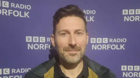 Alex Arnold/BBC A man with brown hair and a grey stubby beard smiles at the camera. There is BBC Radio Norfolk branding behind him.