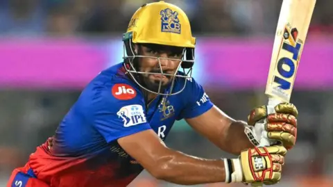Mahipal Lomror in a blue and red shirt, with gold gloves and helmet, hits a shot with a bat facing upwards