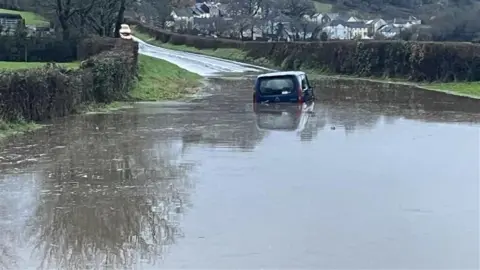 A car is half full of water in a large flood spanning the road. There are houses in the distance.