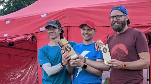 From left to right is Alistair Goulding, Paul McGuiness and
Tom Bradley. Alistair is wearing a blue shirt and a brown cap, Paul is wearing a blue shirt and red cap and Tom is wearing a blue cap and a burgundy T-shirt They are all holding Kubb trophies in their hands.