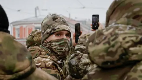 A man wearing camouflage looks into the distance while others around him, also in camo, raise their phones to photograph something