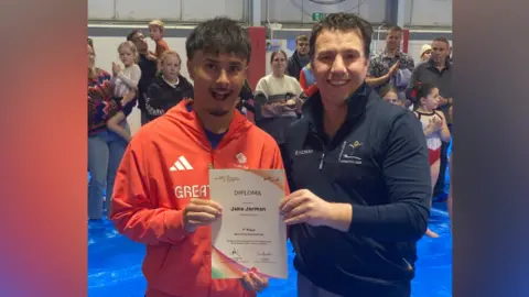 James Burridge/BBC Jake Jarman is stranding on the left in a red Great Britain team hoodie and is smiling at the camera. He is holding a white certificate with a man on the right, who is wearing a black jacket. 