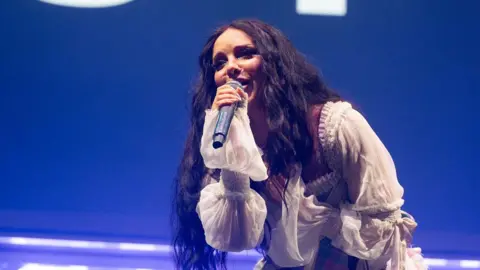 Jade Thirwall performing at the Big Weekend. She has long dark hair and is wearing a white dress. 