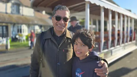 Pawan Adhikari and his son are standing together in front of a tram carriage on a sunny day. Pawan is wearing sunglasses. They both have short, wavy dark hair.