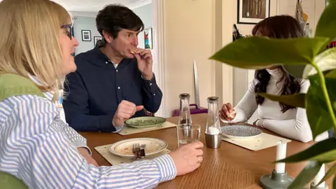 Shula, whose face is obscured by a plant, is pictured finishing lunch at a wooden dining table, with her hosts, Beverley and Mark. She is wearing a white high-neck top and eating bread. Beverley is wearing a blue and white striped top. Mark is wearing a navy top and taking a bite of bread.