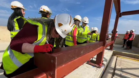 Several people wearing white hard hats and high viz vests are standing on the roof of an unfinished building. One of them is writing on a maroon girder with a pen.