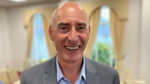 BBC Deputy Steve Williams - A man with a bald head smiling at the camera. He is wearing a grey suit and a blue shirt. 