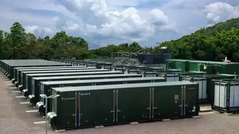 A row of large battery storage containers, which resemble lorry containers but a bit longer, are pictured on an industrial site, with trees and blue sky in the background.