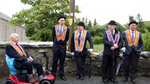 Five men are side by side in front of a stone wall. Each of them are wearing orange and purple sashes around their necks, dark blazers and bowler hats. 
The man on the far left is sitting in a red mobility scooter