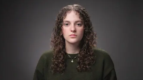 A woman looks at the camera. She has long curly hair and is wearing a dark green jumper and a Star of David on a chain around her neck
