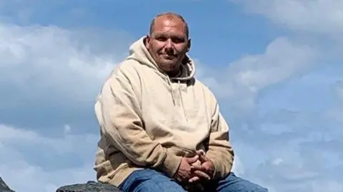 Charlie O'Sullivan in a cream coloured hoody and blue jeans sitting down on some rocks. He is looking directly at the camera against the backdrop of broken clouds.