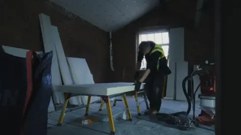 Aiden Joseph A workman stands over a piece of wood with a saw in his hand. The room is other wise bare and dusty
