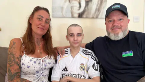 Leo sits in between his mother and father on a sofa in the living room of their house. His mother wears a white strappy floral dress and his father wears a Severn Trent branded navy cap and t shirt. His mother has long brown hair and his father has a grey beard. The wall behind them is a magnolia colour and there is a photo of an elephant behind them on the wall.