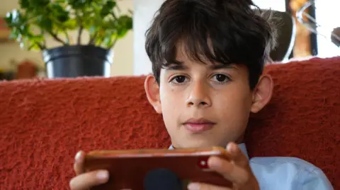 A schoolboy with floppy black hair sits on the sofa in his blue school shirt and tie. He is looking into the camera and holding a smartphone out in front of him.
