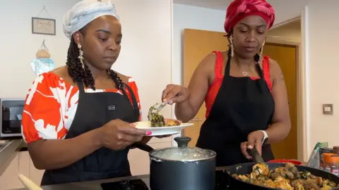 Nyamwererei and Tendai are wearing black aprons stand at a stovetop in a modern kitchen. Nyamwererei holds a plate while Tendai uses a spoon to serve a cooked dish from a frying pan; a pot with a lid sits on the hob nearby.