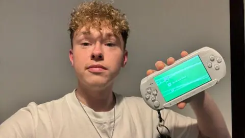 Kyle is indoors holding up a white PlayStation Portable (PSP) console with a keychain attached.