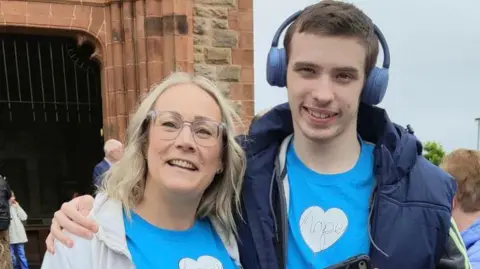 Caroline Campbell A smiling Caroline Campbell is embraced by her teenage son Oscar as they both stand outside the Guildhall in Derry. They are both wearing blue t-shirts, Caroline is wearing a grey hoodie while Oscar, who is also smiling, is wearing a navy jacket and headphones on his head