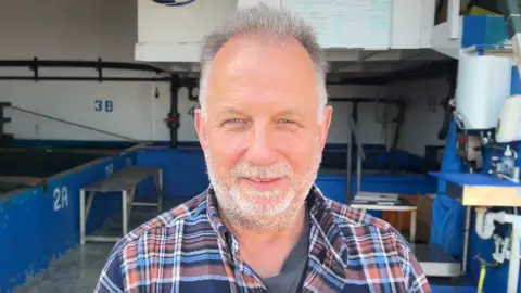 Peter Tarrant has short grey hair and a trimmed beard, wearing a brown and blue plaid shirt, standing in an industrial workspace with blue tanks, pipes, and numbered stations visible in the background.