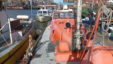 Jemma Sworder An orange lifeboat, in poor condition with chipped and faded paint, is docked in a harbour.