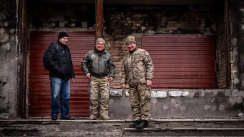 BBC / Claire Jude Press Maksym (L) Volodymyr (C) and 'the colonel' (R) stand in front of their bombed out office for local volunteers