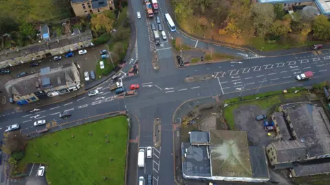 Bradford Council Aerial view of a busy road intersection in an urban area. Multiple lanes with marked turning arrows and central traffic islands are visible. Several cars and buses are traveling in different directions.