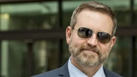 Roger Giese looks at the camera in a photograph from 2018. He wears sunglasses and a suit and has light brown hair and a beard.