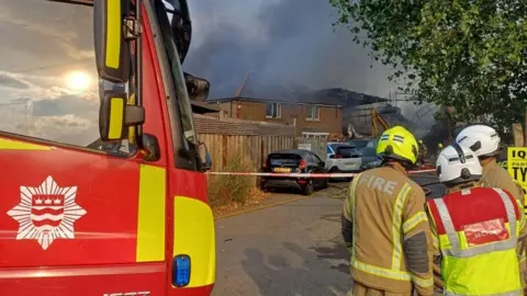 Firefighters stand beside a London Fire Brigade engine as thick black smoke rises from a large fire behind nearby buildings in Mitcham. Emergency crews have cordoned off the area while tackling the blaze at a workshop and scaffolding yard.