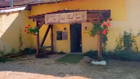 Claire and Neal Hensley A rustic-looking French pub with a wooden porch and the sign The Boar's Head Inn across the top. The wall of the pub is yellow and there are two hanging baskets of red flowers on the porch strutts. There is gravel leading up to a green mat laid in front of a plain black door