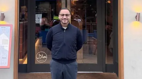 Otto Mellouki Otto Mellouki, who wears dark glasses, jacket and trousers, smiles as he poses outside his restaurant. The entrance is behind with a Halal sign on the glass door and outdoor menu on the left.