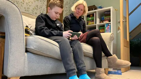 BBC A young boy in a black top looks down at a phone while a faired haired woman looks at him.