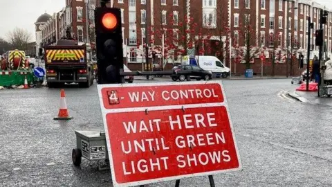 A temporary red roadworks sign with white writing which says: "Four way control. Wait here until green light shows". A temporary traffic light is showing red and in the background there are two highway maintenance vehicles.