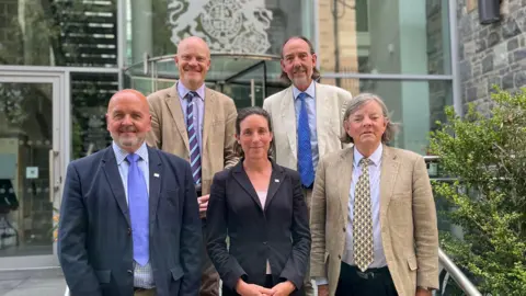 The five original members of this term's Policy and Resources Committee standing in front of Guernsey's Royal Court. 

Standing in the back row is Deputy Gavin St Pier on the left, who is a bald man weariung a brown suit, purple shirt and a purple and turquoise tie. Next to him is Deputy Steve Falla, who has mid-length brown hair and is wearing a cream suit jacket, blue shirt and blue tie. 

On the front row is, on the far left, Deputy Jonathan Le Tocq, a bald man with a grey beard and a blue jacket, blue shirt and blue tie. In the middle is Deputy Lindsay de Sausmarez, she has black mid-length hair and a black jacket on top of a pink blouse. She's wearing a Guernsey pin badge on her left lapel. On the far right is Deputy Charles Parkinson, a man with mid-length grey hair, a brown suit and a blue shirt with a yellow and blue checked tie. 