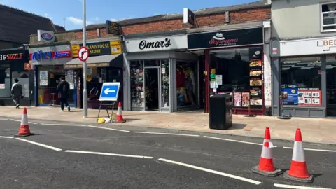 Nicola Haseler/BBC Midland Road in Bedford, showing traffic cones and shops along the road. There are road signs and two people walking along.
