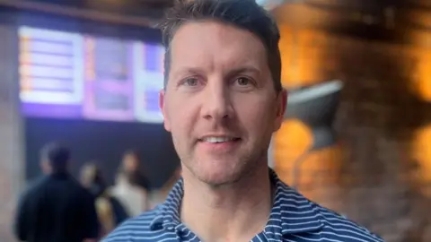 A man with brown eye smiles into the camera wearing a blue and white striped t-shirt with an open collar. He is standing in front of a blurred crowd ordering from a menu above the cashier.