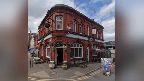 Google The pub stands on the corner of two streets with two barrels outside and some benches. It is a red brick building.
