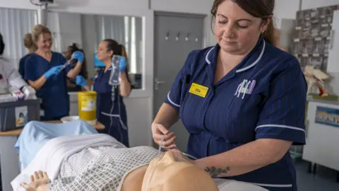 A nurse in blue uniform attaching tubes to a clinical dummy with other nurses holding equipment in the background